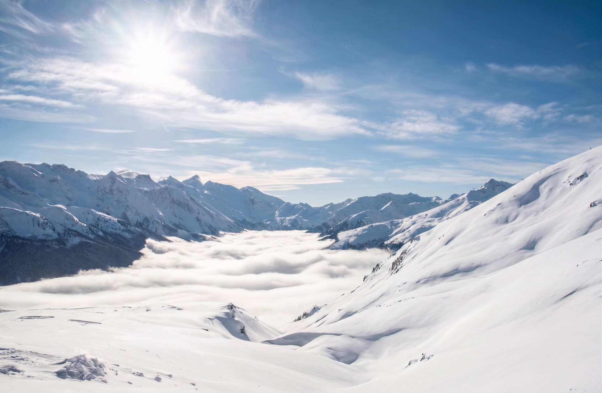 Snow-covered alpine peaks above a valley filled with low cloud, lit by bright winter sun.